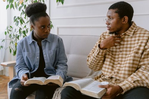 Free Two adults sitting on a sofa, engaged in conversation while holding books, indoors. Stock Photo