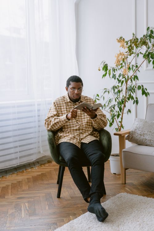 Free A man sitting in a chair reading a book by the window in a cozy living room setting. Stock Photo