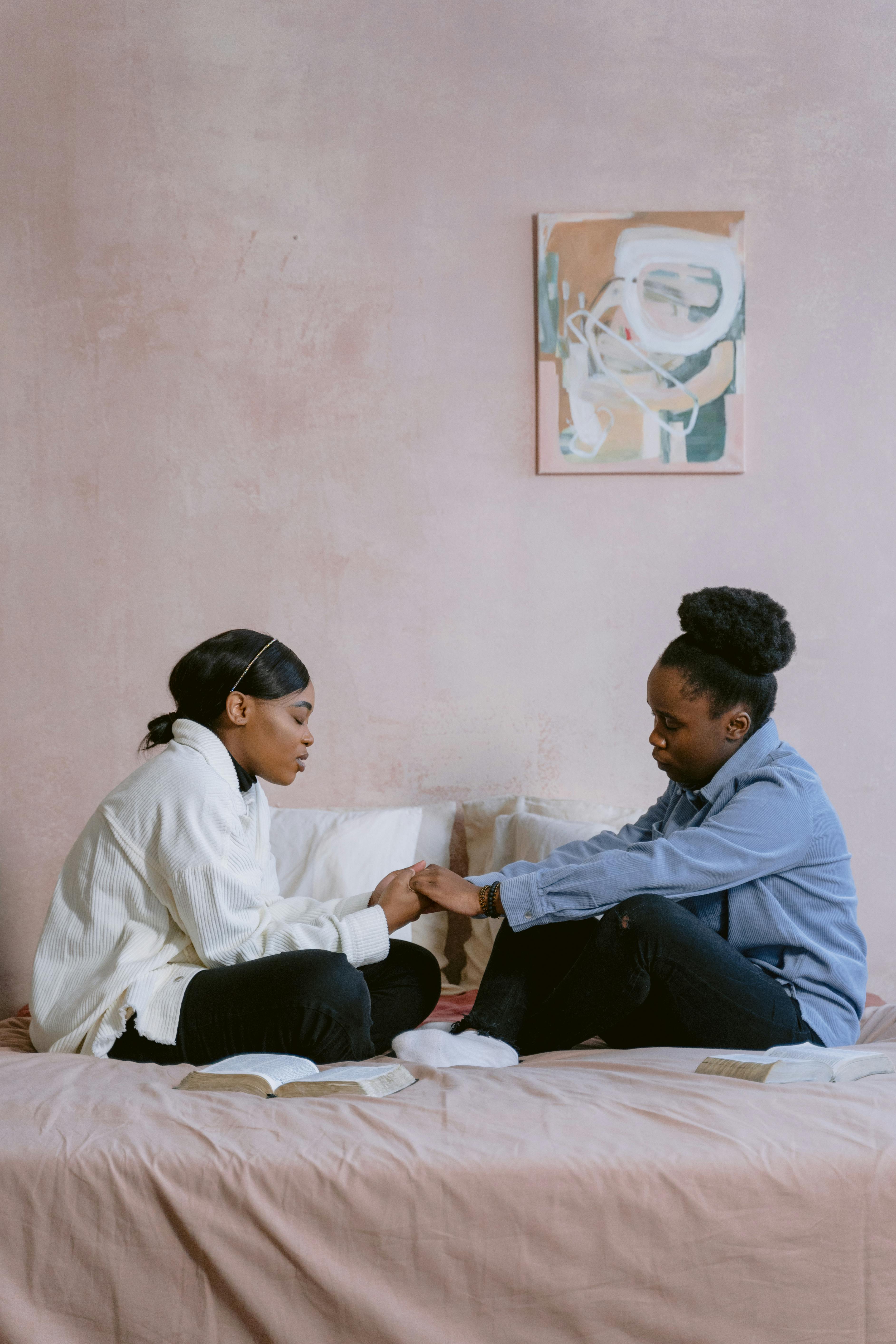 Two women holding hands and praying on a bed in a serene bedroom, symbolizing faith and connection.