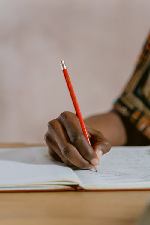 Person Holding Red Pencil Writing on Notebook