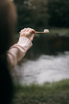 A person holds a bamboo toothbrush in front of a serene outdoor setting by the water.