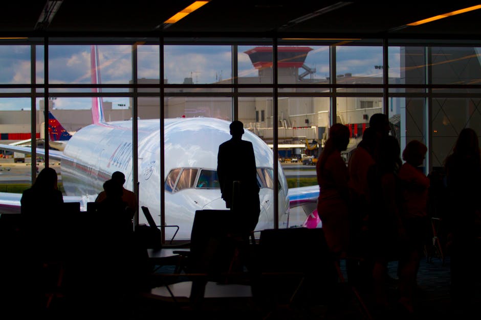 Photo by Terrence Bowen Silhouettes of travelers waiting at an airport terminal with an airplane visible through the window.