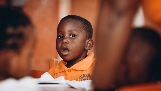 A young African American boy in an orange polo shirt exhibits a curious expression during an indoor setting.