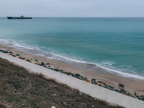 Picturesque scenery of ship sailing on wavy sea with foamy waves rolling on sandy coast against cloudy sky