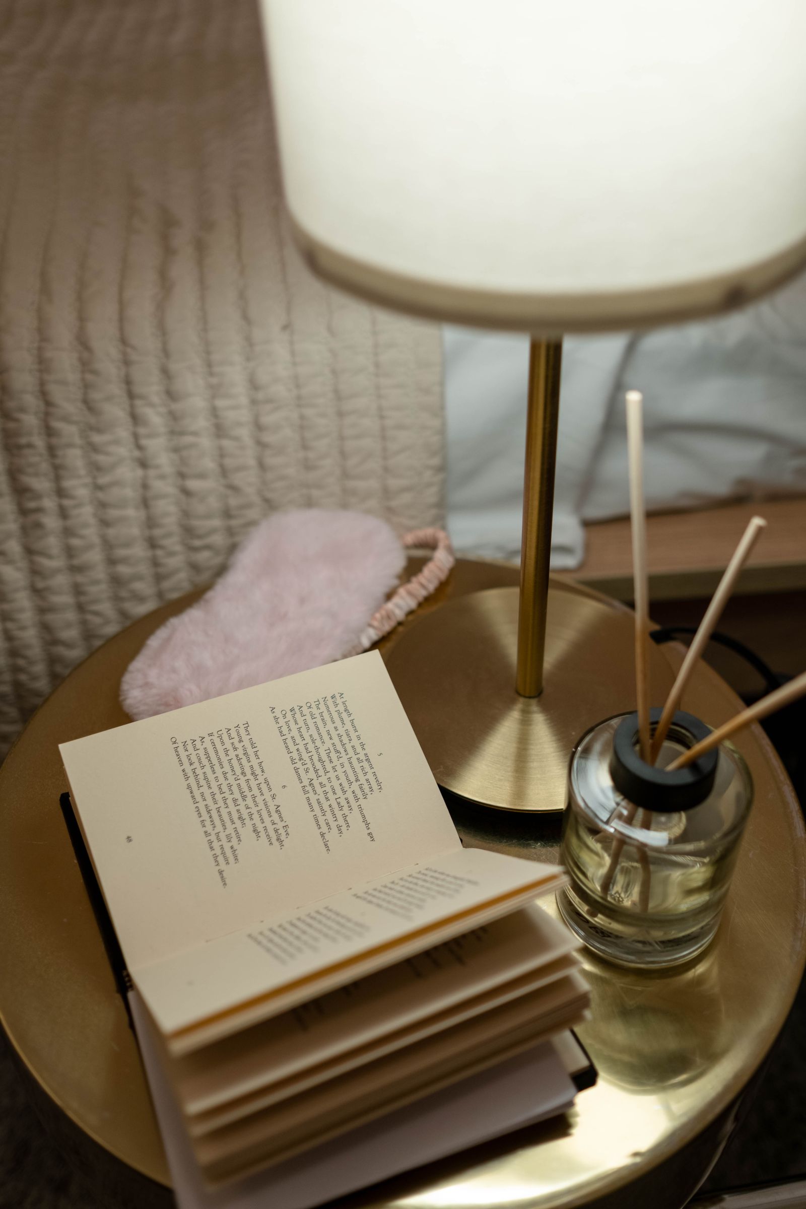 A bedside table with an open book, a warm brass lamp and an eye mask