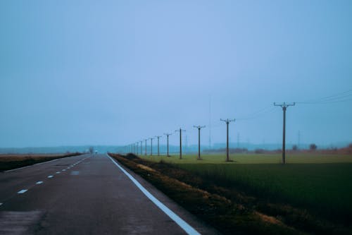Straight asphalt roadway against row of electric poles in field under blue sky in daylight