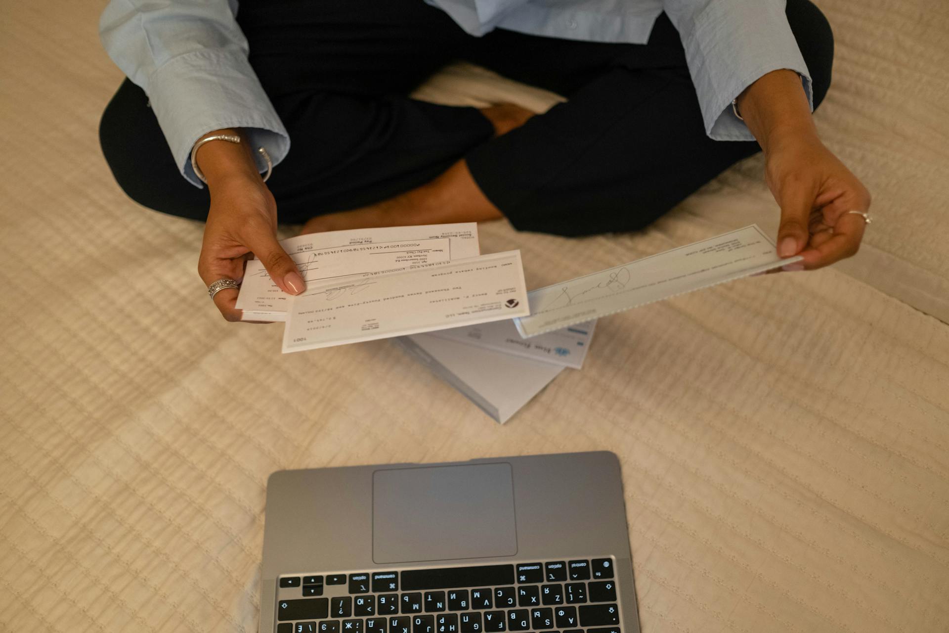 Close-up of hands holding cheques beside a laptop indoors for financial tasks.