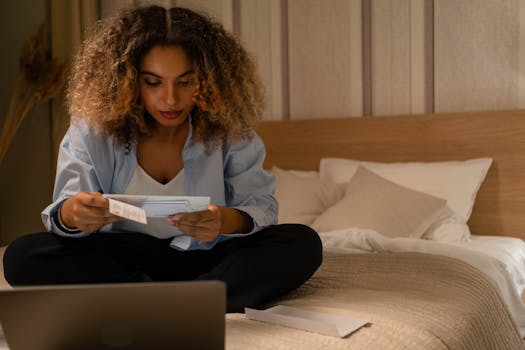 Woman sitting on bed reading documents with laptop, highlighting home office setting.