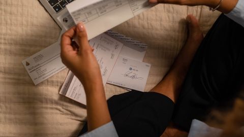 Woman holding checks while managing finances on a laptop, showing online banking on the screen.