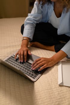 A woman sits cross-legged on a bed, typing on a laptop, representing work-from-home lifestyle.