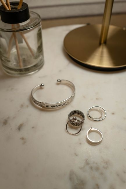 Close-up of silver jewelry pieces on a marble surface with a glass diffuser and lamp.