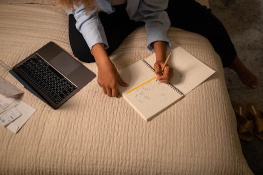 Casual home workspace with a woman writing in a planner on a bed with a laptop.