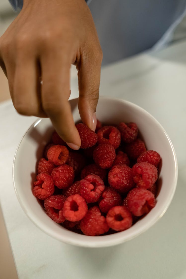 A Person's Hand Getting A Raspberry From A Bowl