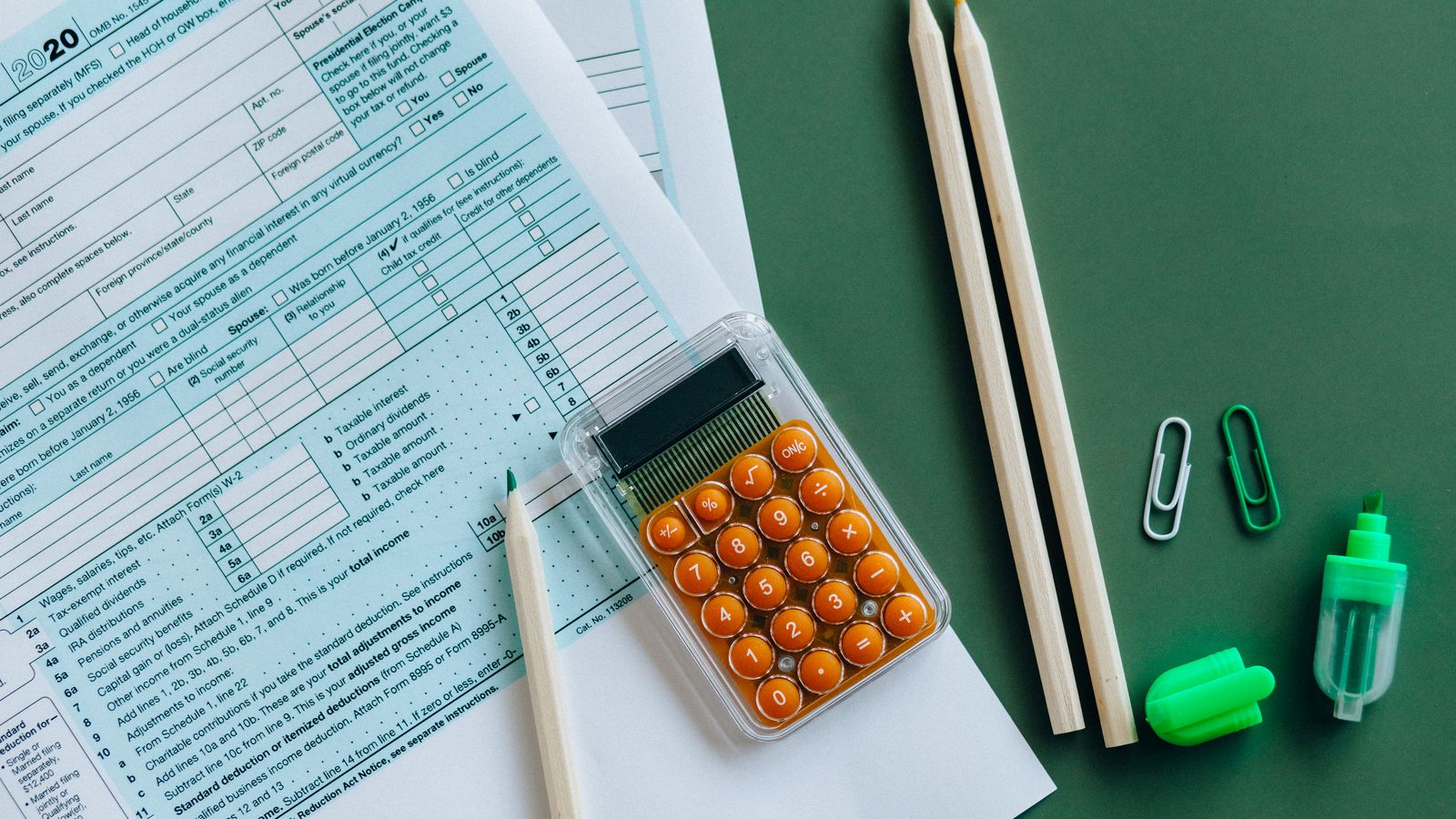Flat lay of tax forms, calculator, pencils, and clips on green surface, ideal for finance or accounting themes.
