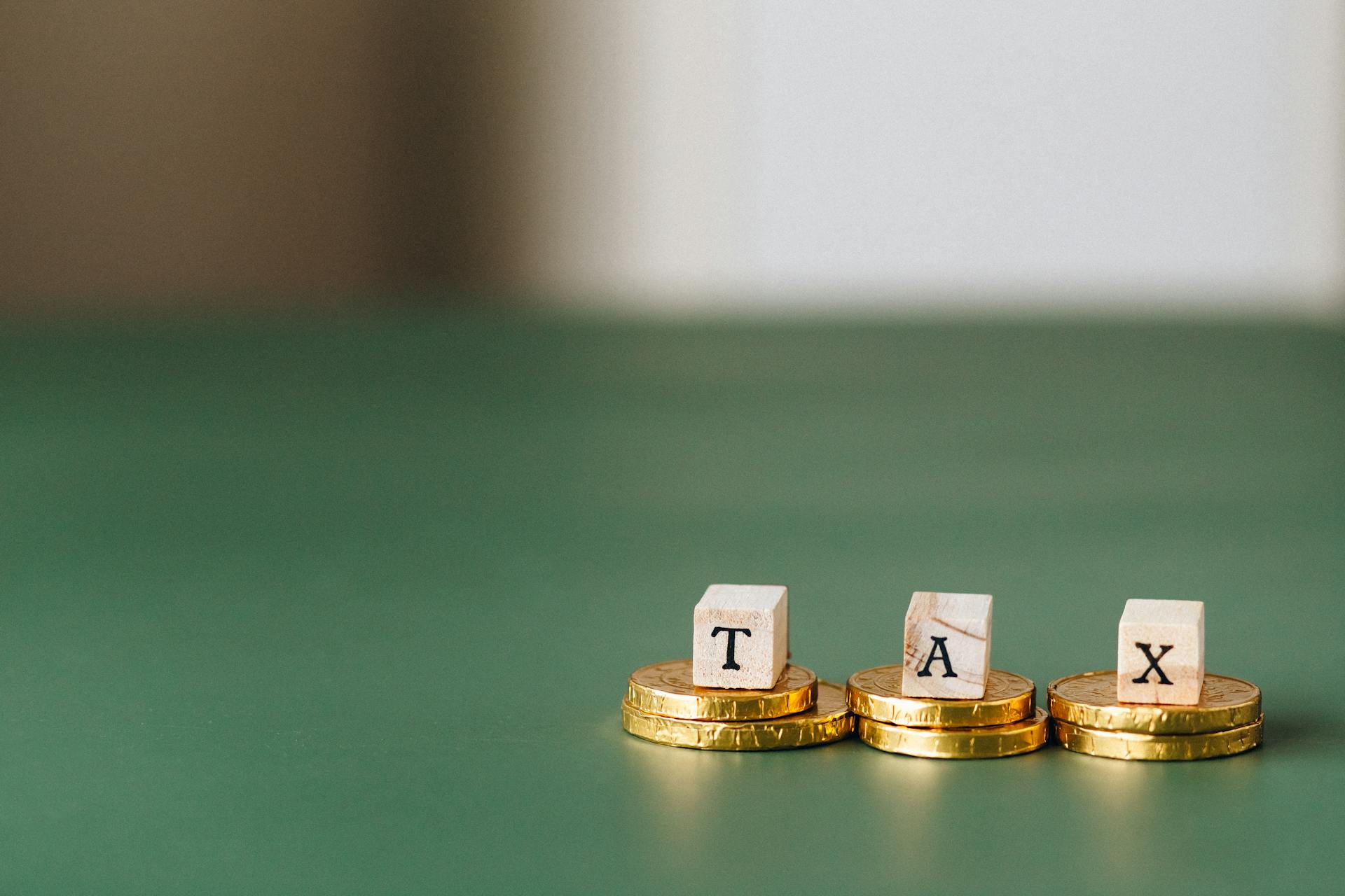 Stacked gold coins with wooden blocks spelling 'TAX' on a green backdrop.