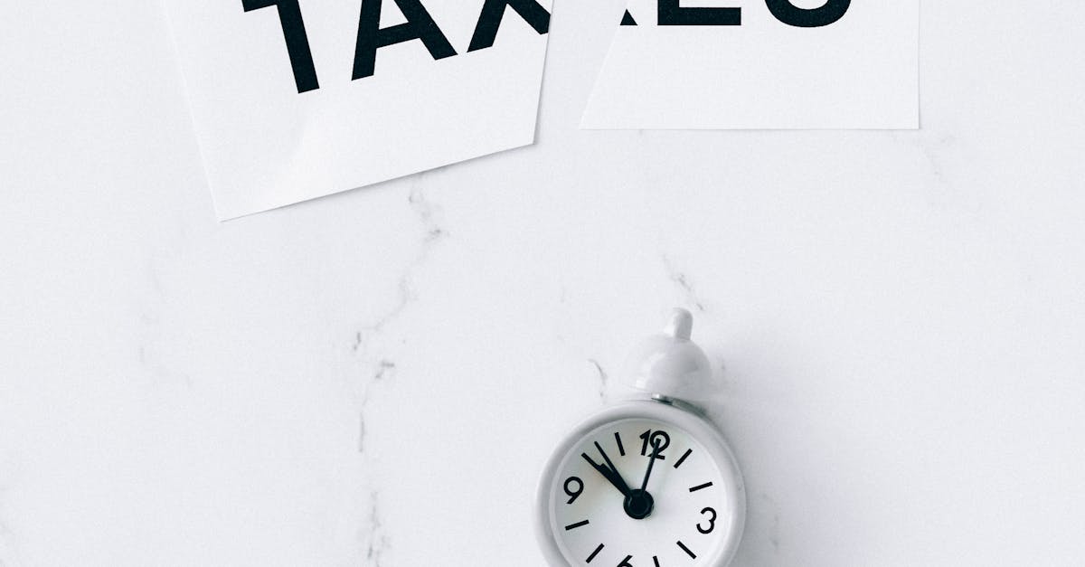 Photo by Nataliya Vaitkevich A minimalist composition featuring a small white clock and torn paper with the word 'Taxes' on a marble surface.