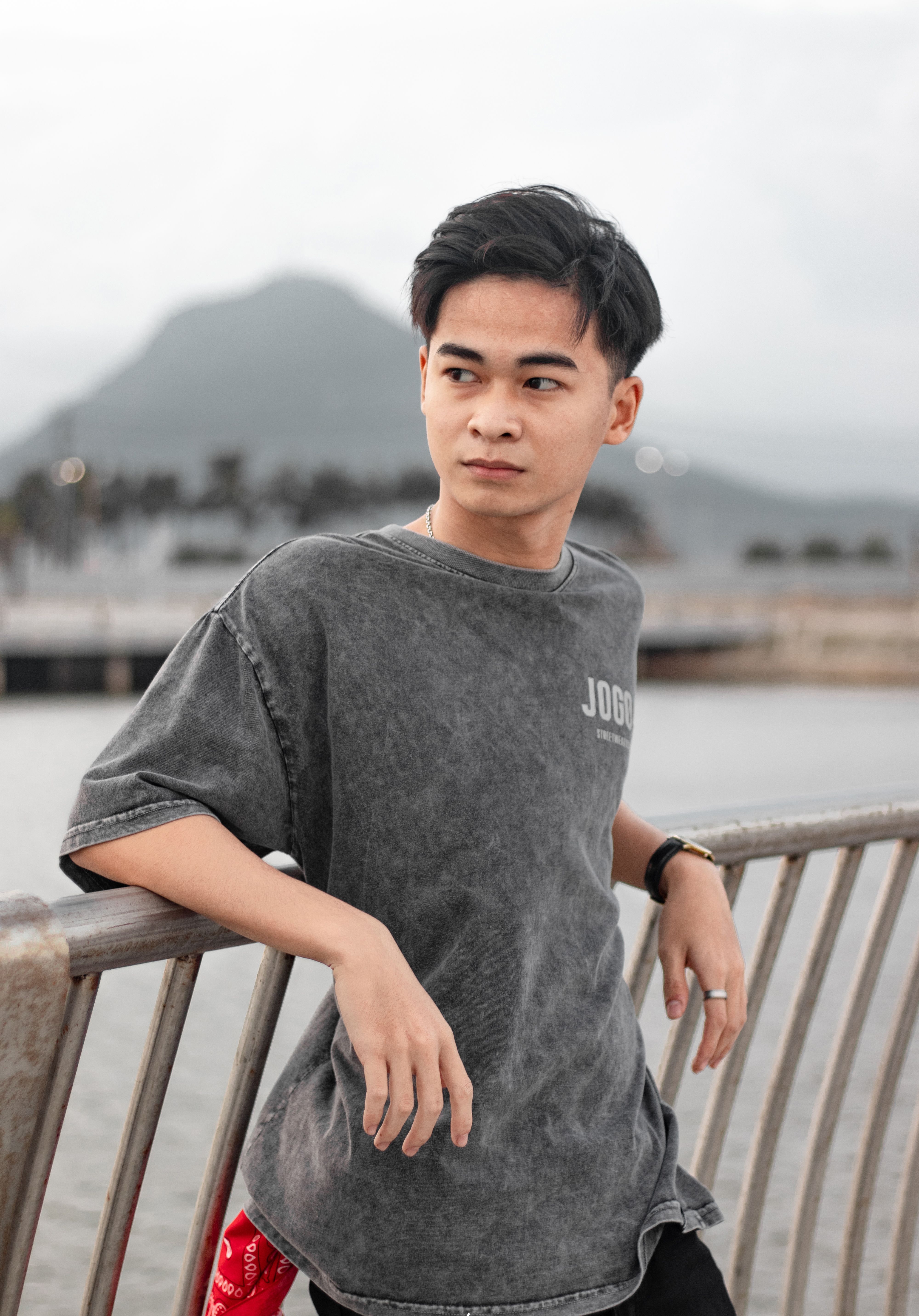 A young man leaning on a bridge railing, looking away with a distant hill in the background.