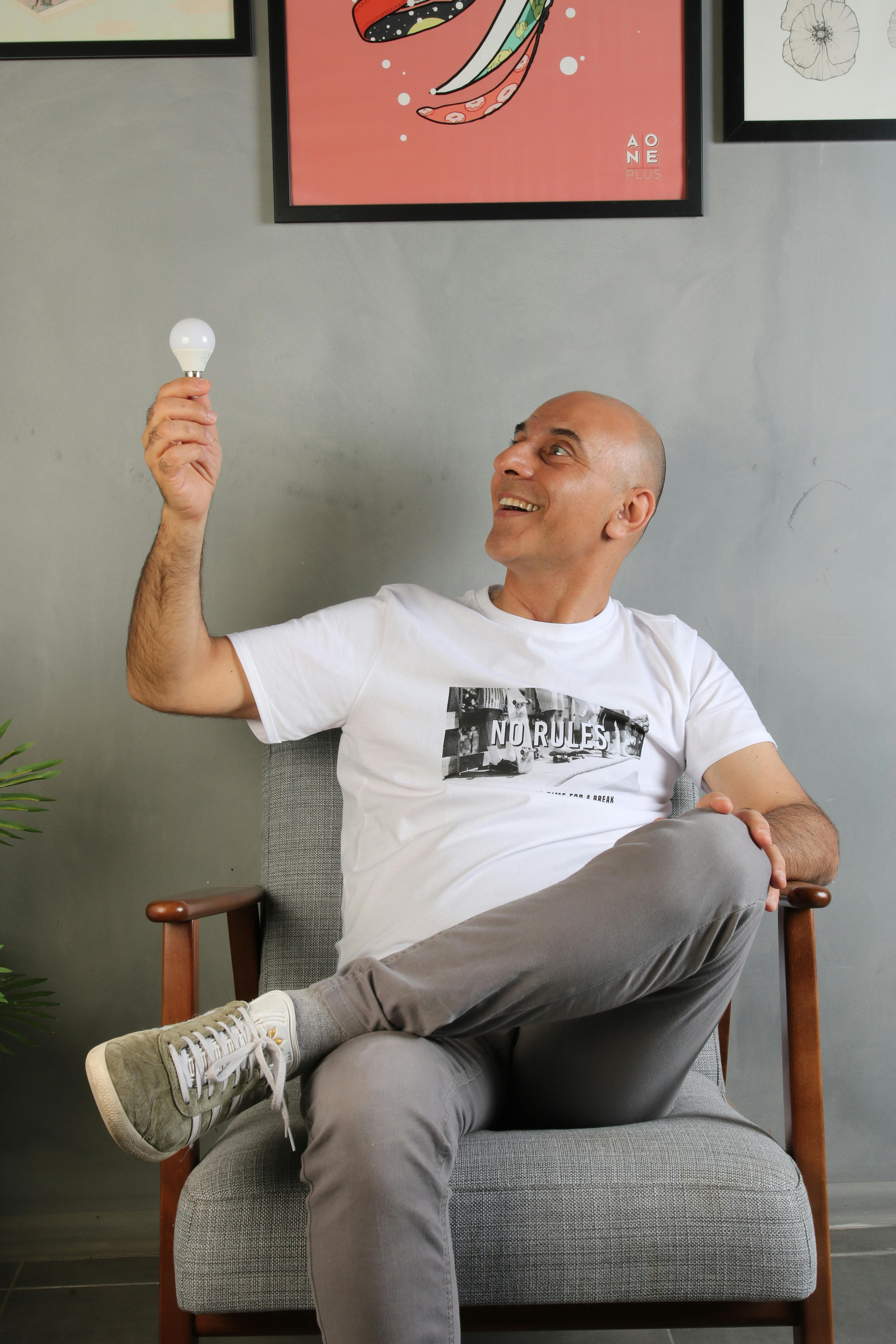 Bald man smiling while holding a light bulb sitting on an armchair indoors.