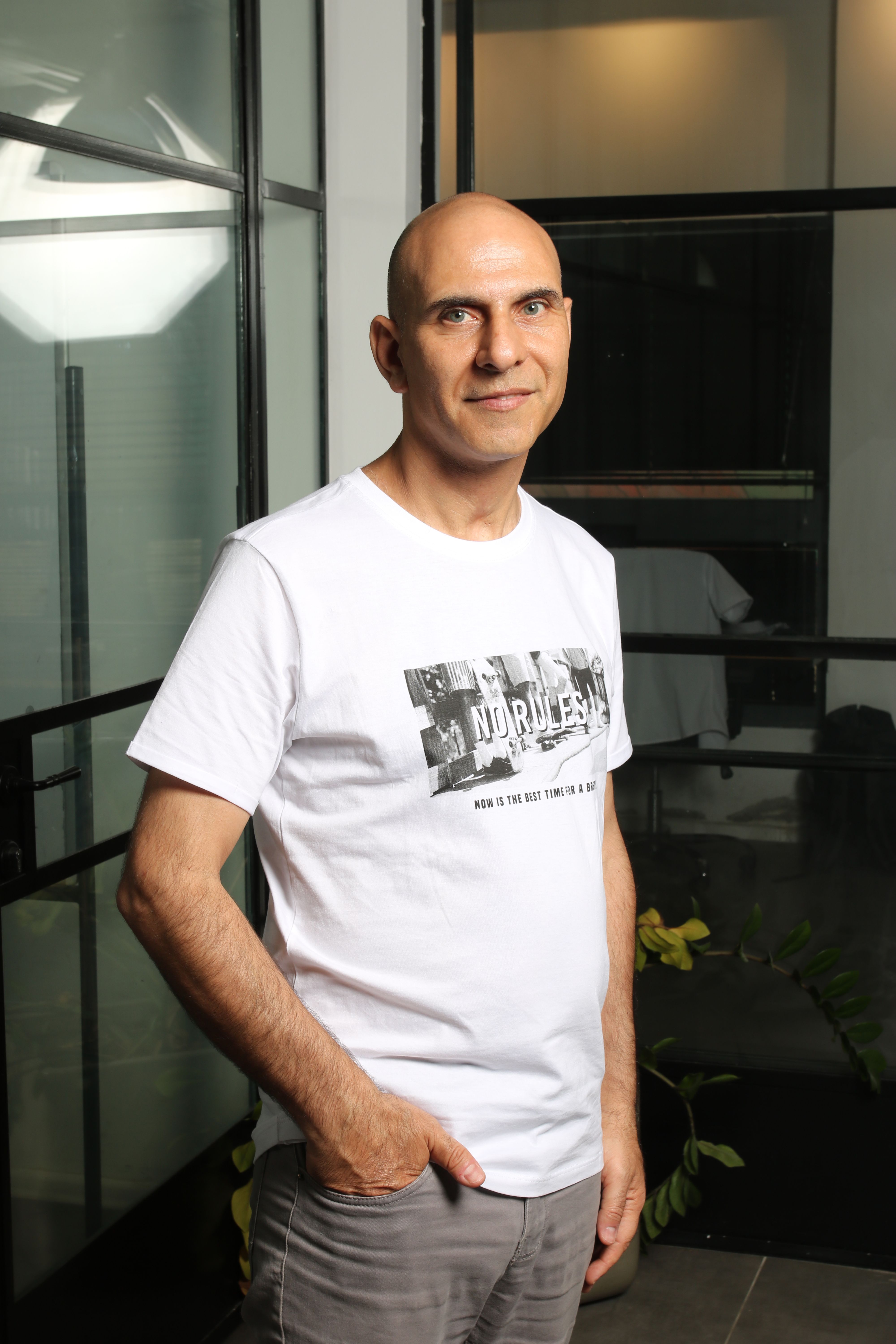 Man with a bald head posing indoors, wearing a white shirt, against a modern background.