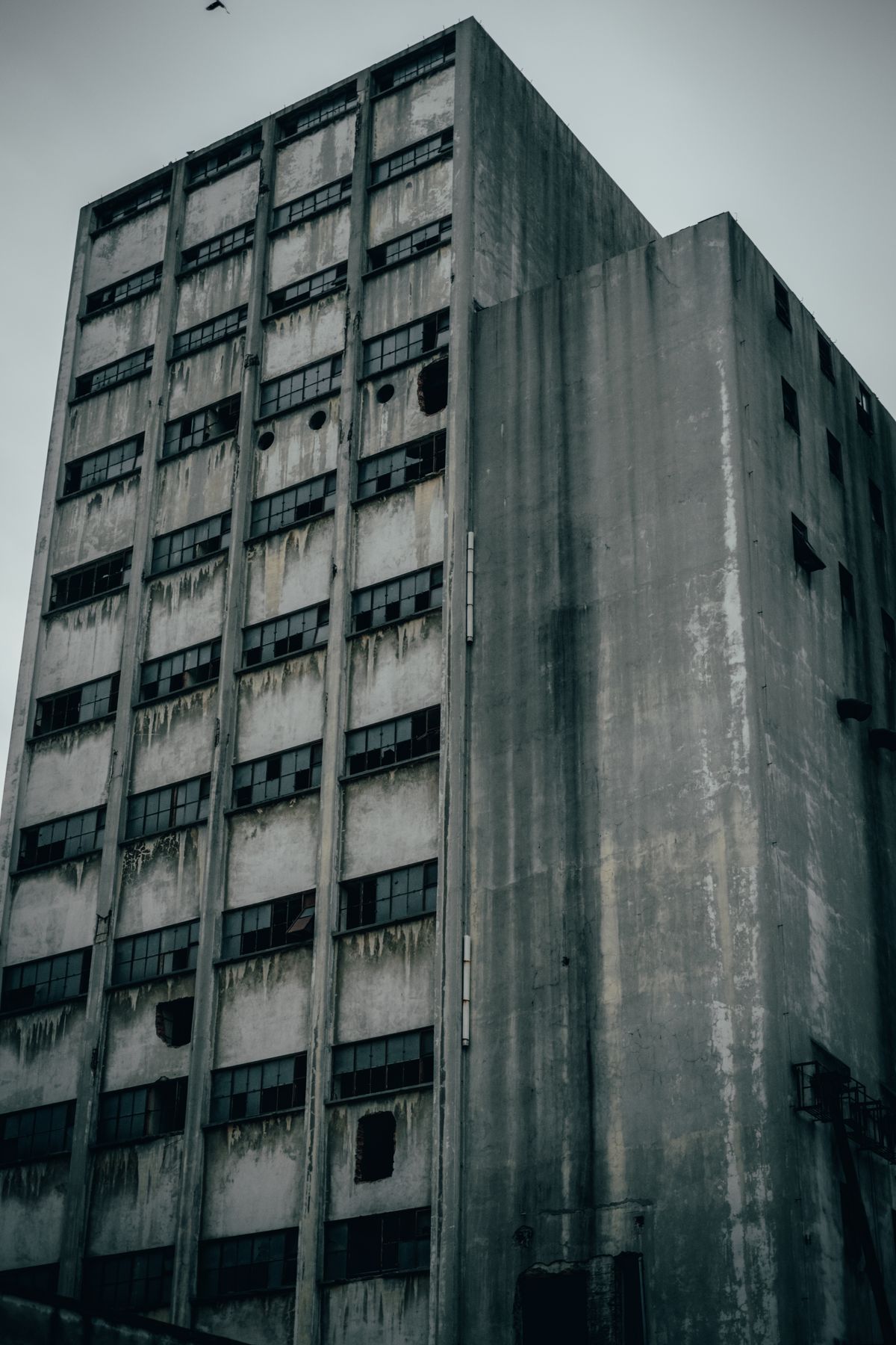 A multistory cement building with corrosion and broken windows in a neglected district, similar to Hashima ruins
