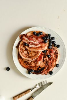Aerial view of pancakes topped with blueberries and crispy bacon on a white plate.