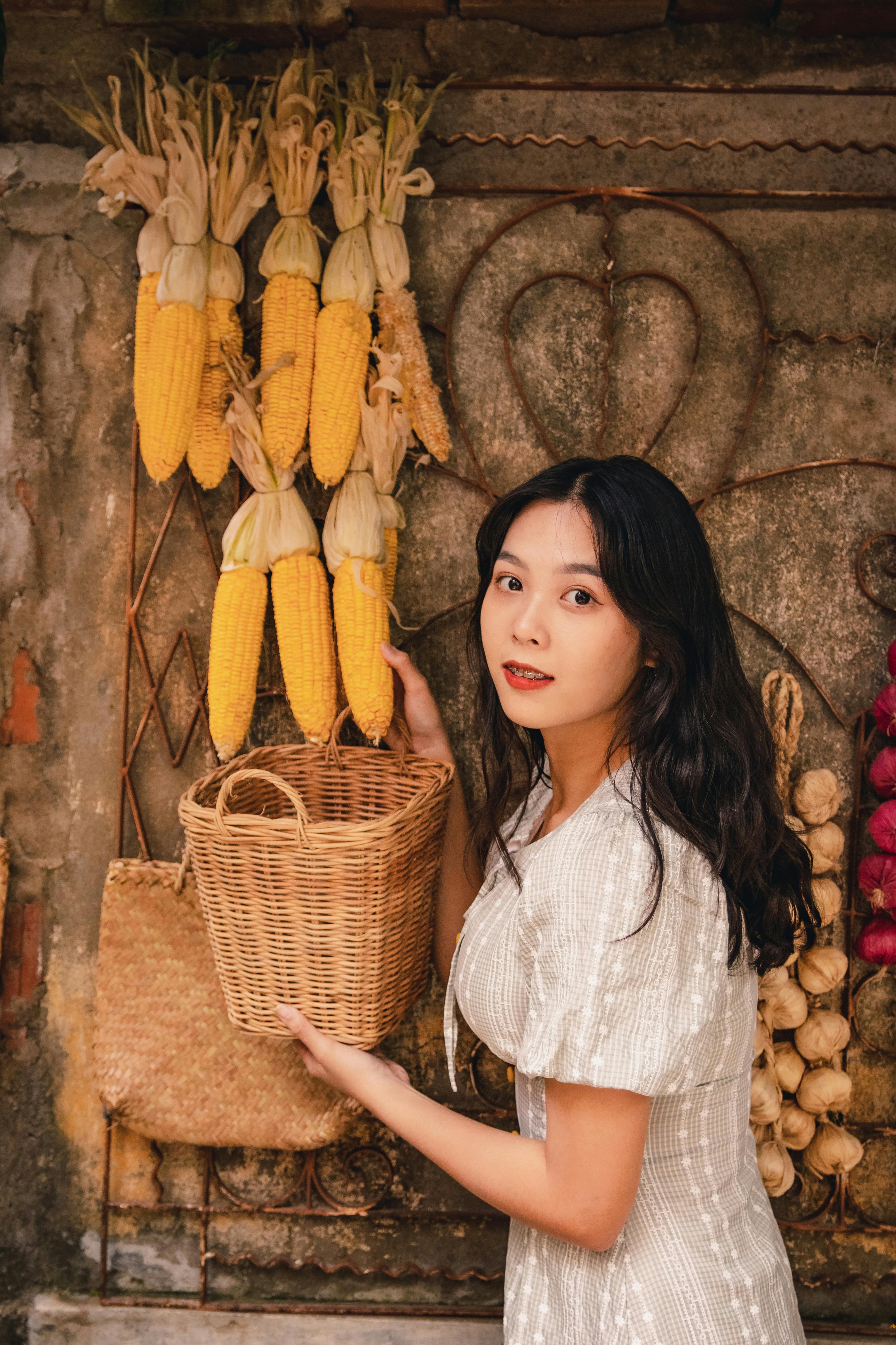 Portrait of an Asian woman holding a wicker basket near hanging corns in a market setting.