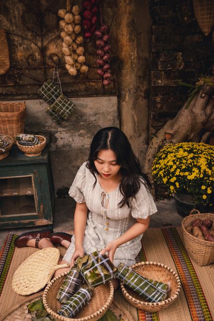 A woman sits with baskets of banh chung and vegetables in a rustic setting.