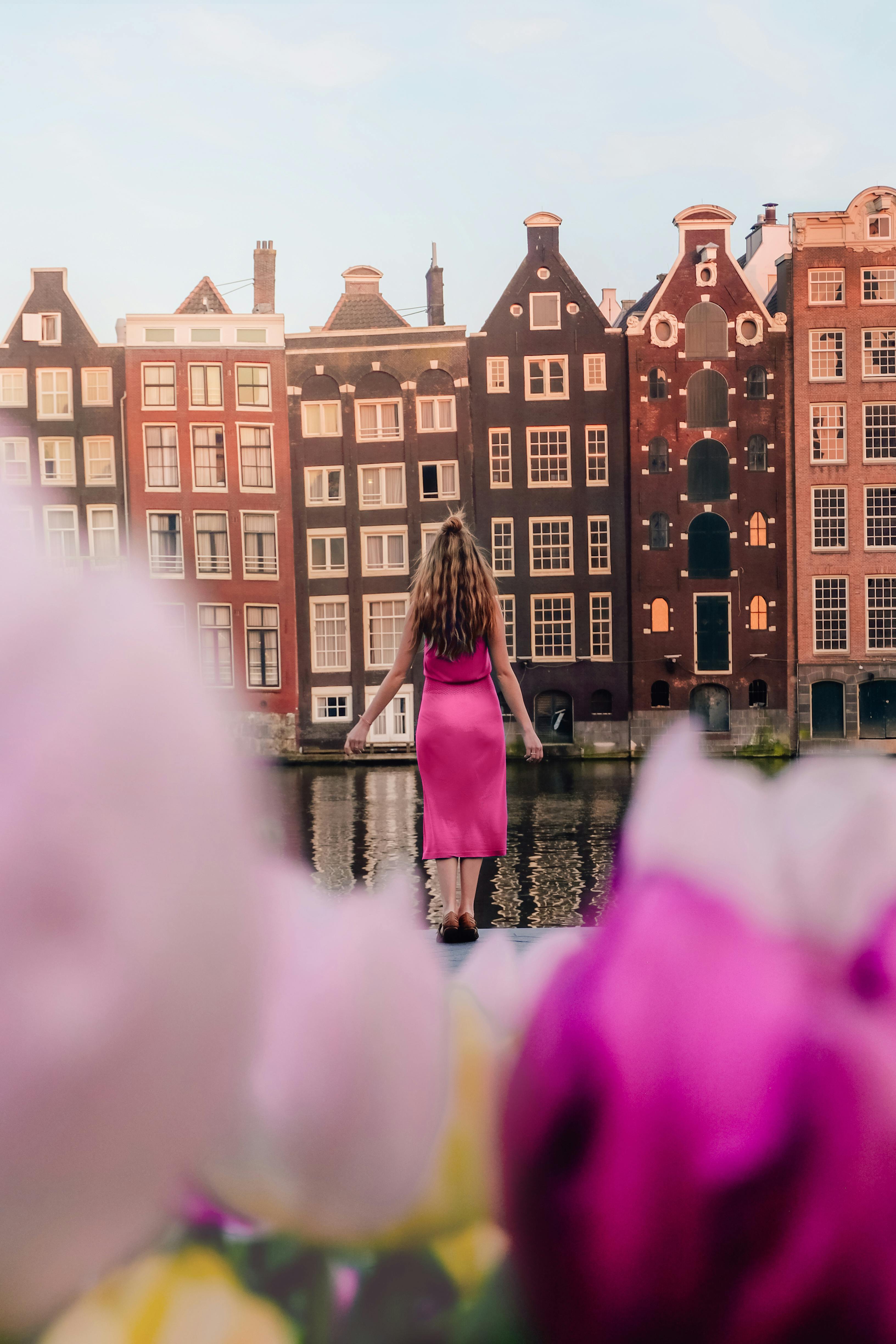 A woman in a pink dress stands by Amsterdam's traditional canal houses, framed by vibrant flowers.