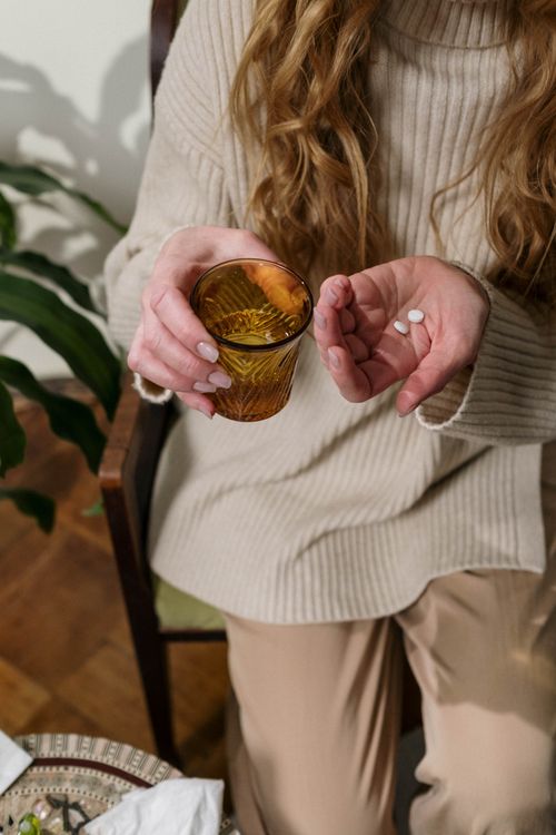 Free A woman sitting indoors holds pills in one hand and a glass of water in the other, implying medication. Stock Photo