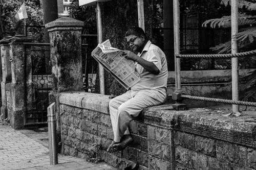 Free Black and white photo of an elderly man reading a newspaper while sitting on a wall in a street in Mumbai. Stock Photo