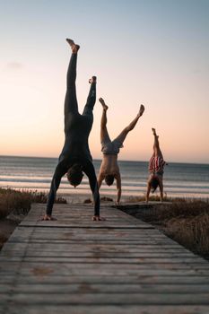 Three people doing handstands on a wooden pathway by the beach during sunset.