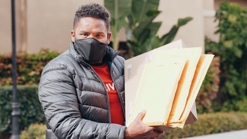 Man in Gray Puffer Jacket with Face Mask Carrying a Box and Envelopes