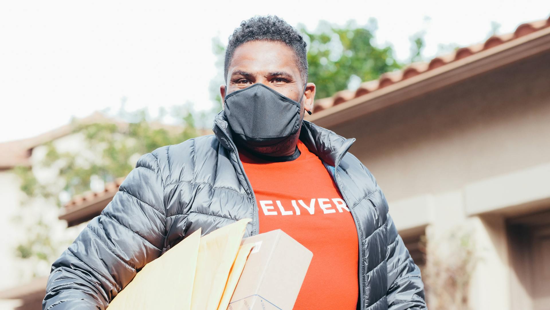 Delivery person wearing a mask holding packages outside a house on a sunny day.