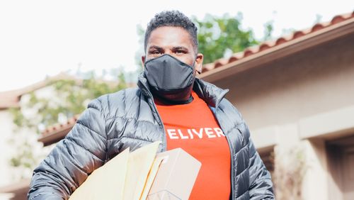 Free Delivery person wearing a mask holding packages outside a house on a sunny day. Stock Photo
