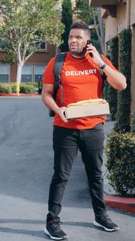 A delivery man in a red shirt stands outside with a package, appearing confused while using his smartphone.