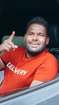 Friendly delivery man in a red shirt smiles through a car window, pointing ahead with confidence.