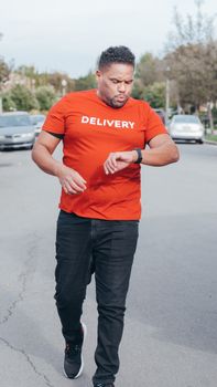 A delivery man in a red shirt checks his watch while walking on a suburban street.