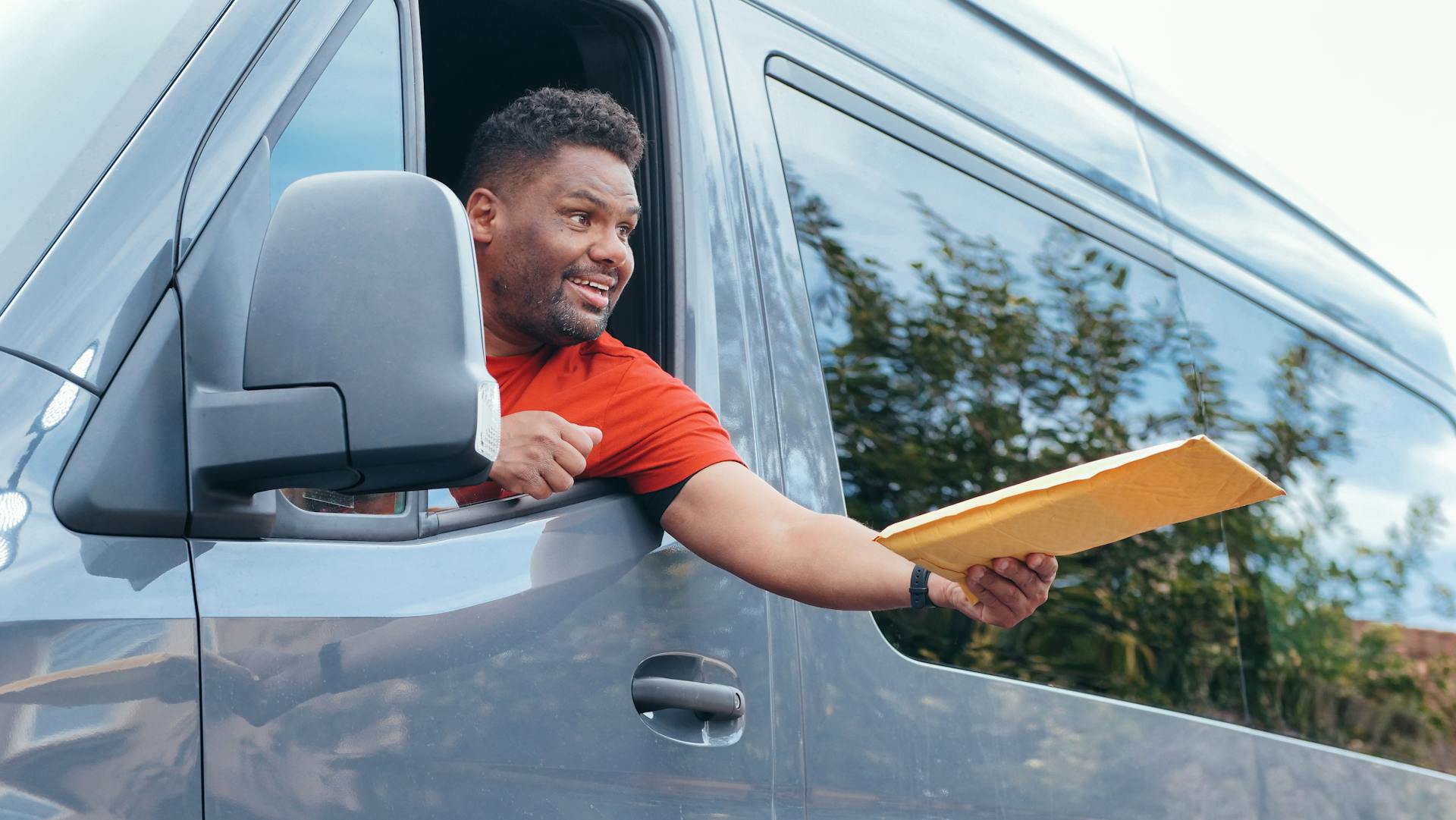 Smiling courier handing out delivery package through van window outdoors.