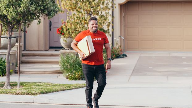 A cheerful deliveryman carrying a package in a suburban neighborhood driveway on a sunny day.