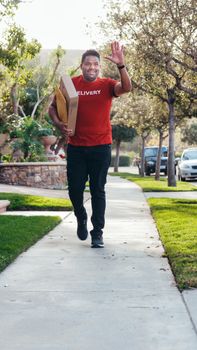 Happy delivery man in red shirt carrying package on a sunny sidewalk, greeting passersby.