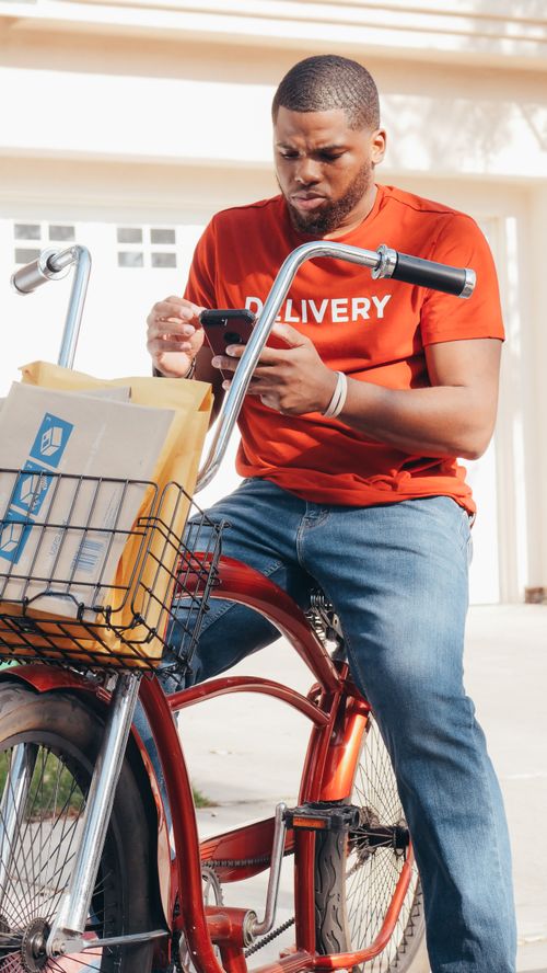 Free A delivery person checks their phone for orders while seated on a bicycle with packages. Stock Photo