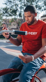 Focused courier in red uniform checks delivery orders on phone and holds packages outdoors.