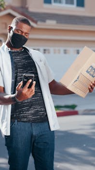 A delivery person checking a smartphone while holding a package in a residential area.