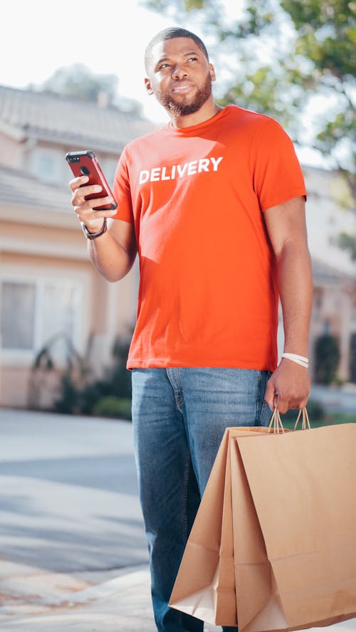 A Man Wearing Delivery Shirt Holding a Cellphone a Brown paper Bags
