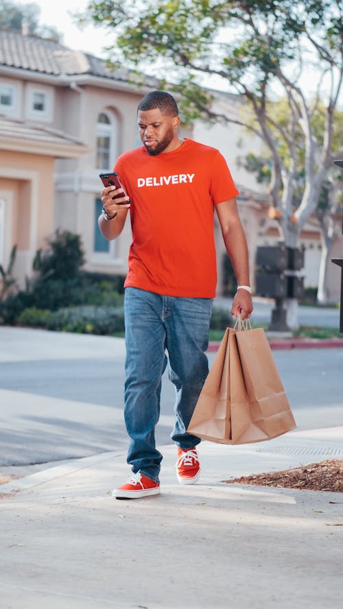 Free Man in Red Crew Neck T-shirt and Blue Denim Jeans Holding Smartphone and Paper Bags Stock Photo
