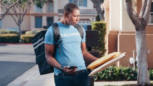 Free Delivery person with smartphone and parcels outside a house in a sunny neighborhood. Stock Photo