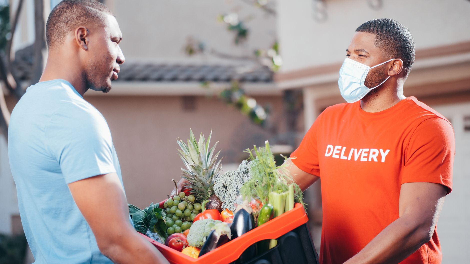 Two men exchanging a box of fresh produce for contactless delivery in a residential area.
