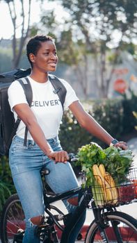 Smiling delivery person on a bicycle with fresh vegetables in the basket outdoors.