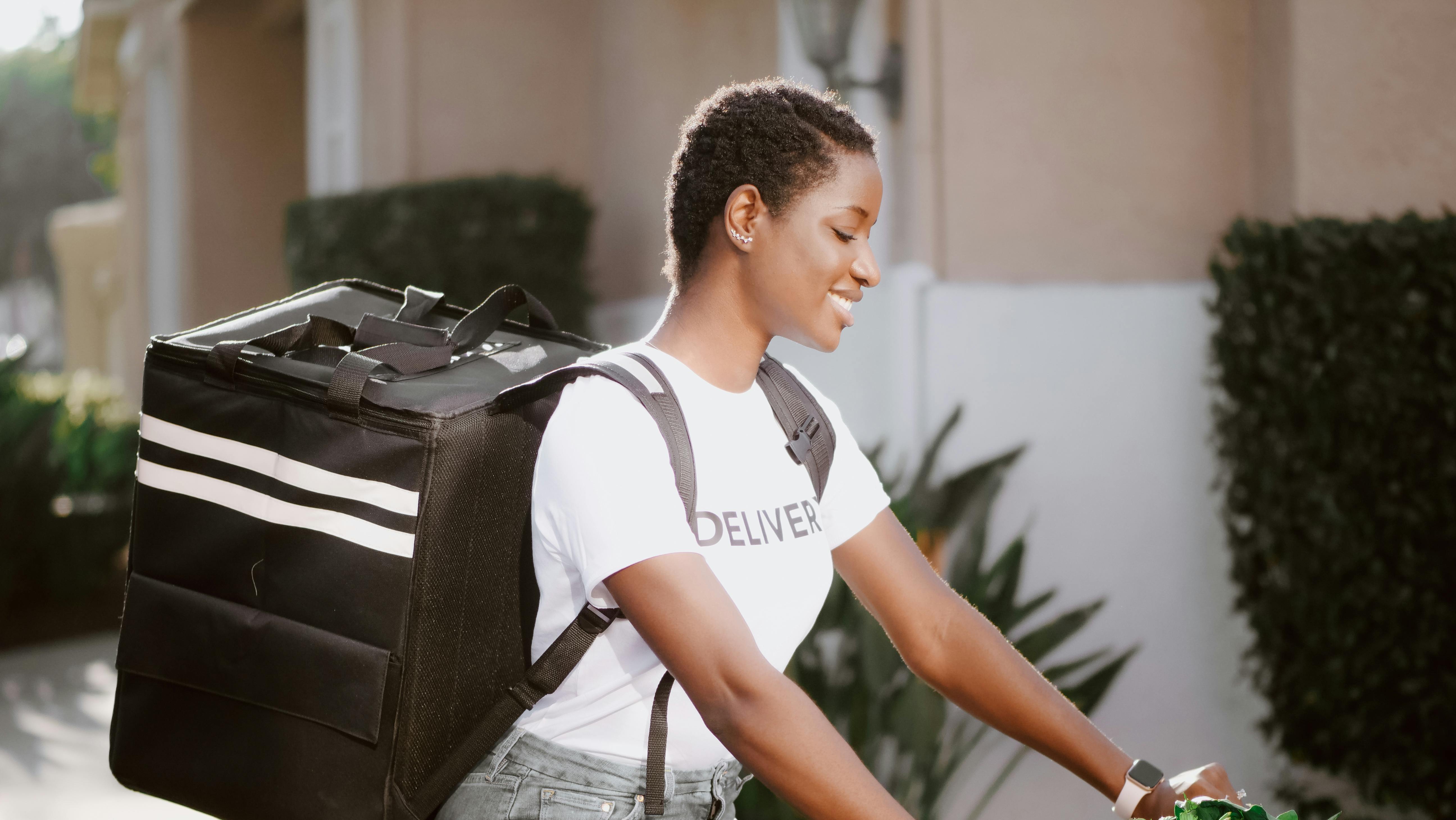 A person with a delivery backpack riding a bicycle outdoors, wearing a white t-shirt, smiling while looking ahead.