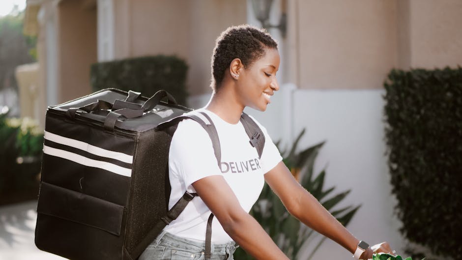 A person with a delivery backpack riding a bicycle outdoors, wearing a white t-shirt, smiling while looking ahead.